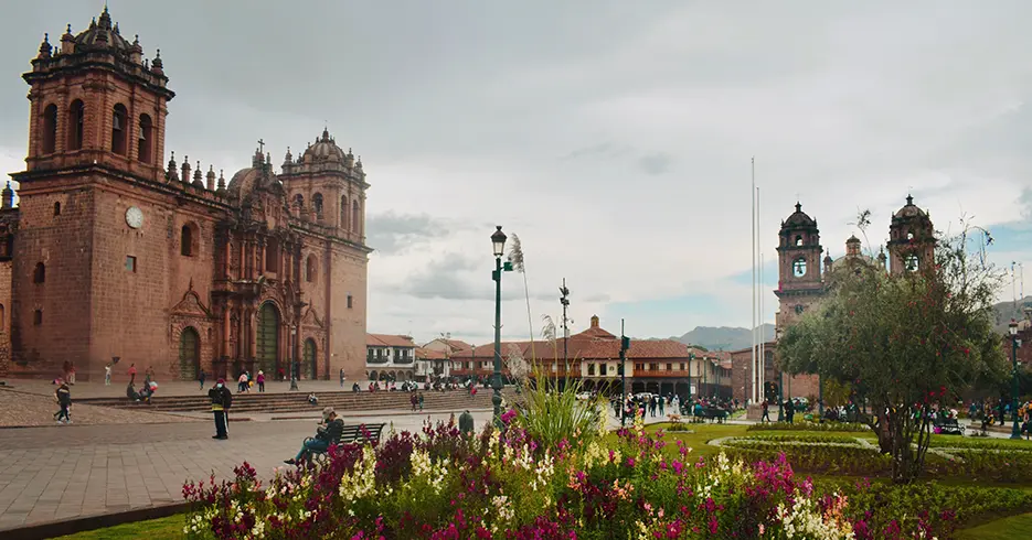 Vista de la flora que se encuentra en la plaza de armas de Cusco, con una vista en la parte izquierda de la catedral del cusco, con un cielo lleno de nubes y se ven bastantes personas alrededor de la plaza