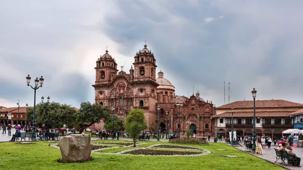 La ciudad del cusco en la plaza de armas con la vista de la iglesia de compañía de Jesús, diferentes personas en la plaza, caminando o sentadas, varios arboles y un jardín verde.