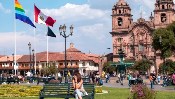 Turista mirando a la izquierda en la plaza de armas de cusco, al fondo se ve la bandera del Perú, y de cusco, también se ve la iglesia de la compañía de Jesús y el inca en la pileta de agua