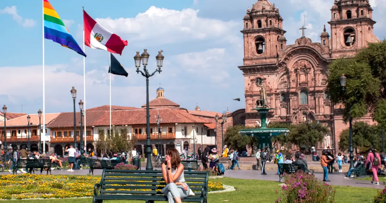Haz Realidad Tu Viaje a Perú en 7 Días 8 Turista mirando a la izquierda en la plaza de armas de cusco, al fondo se ve la bandera del Perú, y de cusco, también se ve la iglesia de la compañía de Jesús y el inca en la pileta de agua