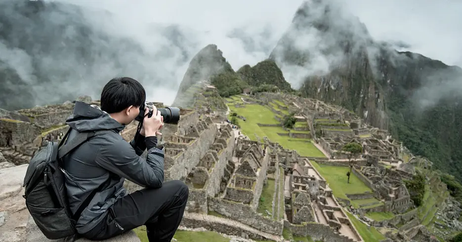 Turista fotografiando Machu Picchu, en la parte baja las construcciones incas y las montañas están cubiertas con niebla