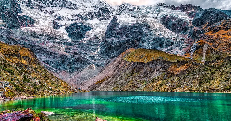 Laguna humantay en un viaje a los Andes en cusco, agua turquesa, verde y azul cristalina, con el nevado humantay de fondo y montañas rocosas con pasto verde