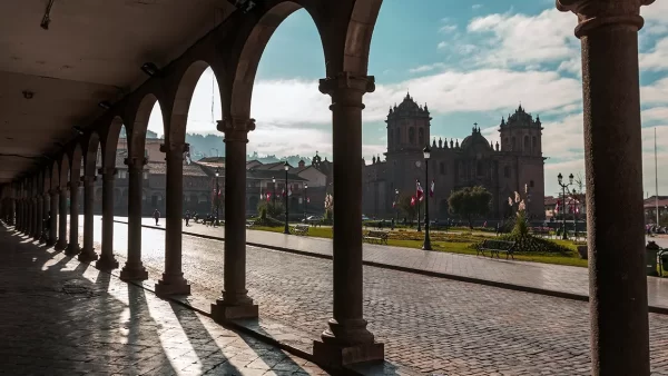 Vista de la plaza de armas y la catedral de Cusco desde los arcos de la plaza, varias banderas de Perú en la plaza y con pasto verde y bancos para sentarse