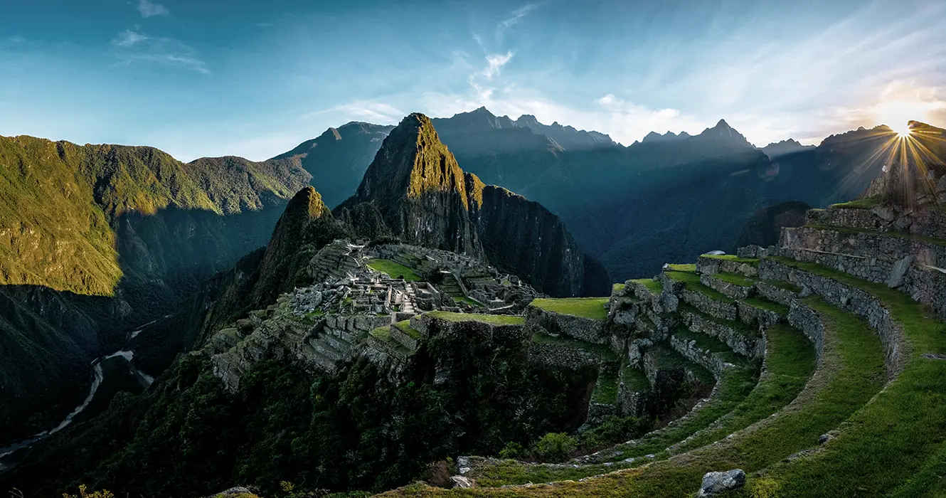 Machu Picchu con un cielo celeste y saliendo el sol, vista de los andenes, las construcciones incas y en la parte baja a la izquierda el río urubamba y muchas montañas rodeando Machu Picchu