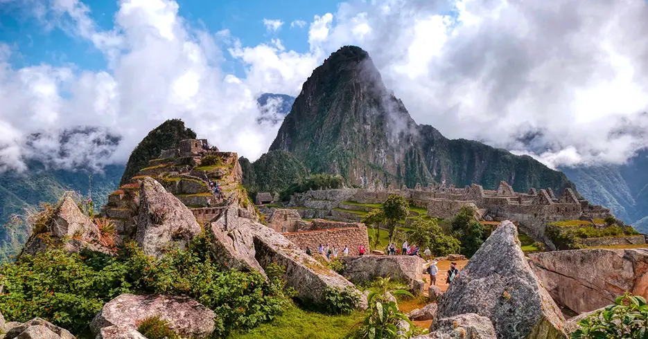 Machu Picchu en cusco, con varias personas, se ven varias piedras y pasto verde y los espacios arqueológicos de fondo en la parte baja, un cielo con varias nubes y una parte el cielo azul