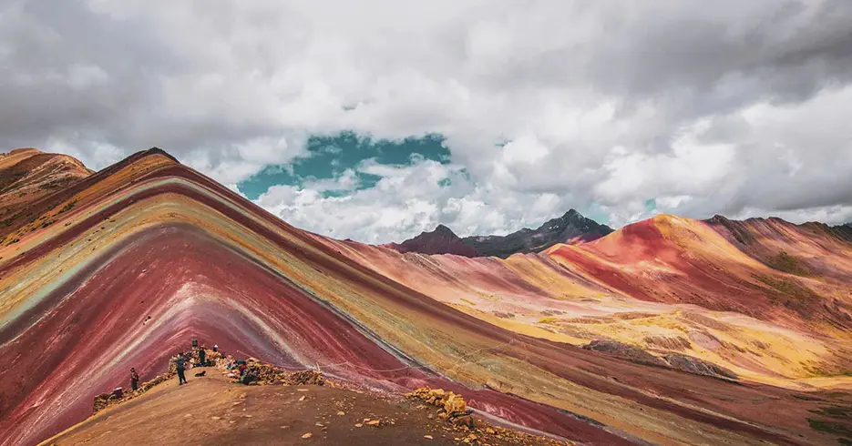 La montaña de los 7 colores en un viaje a los Andes, se ven varios colores, como el rojo, amarillo, naranja y blanco, con un cielo un poco nublado y varias personas en la parte izquierda