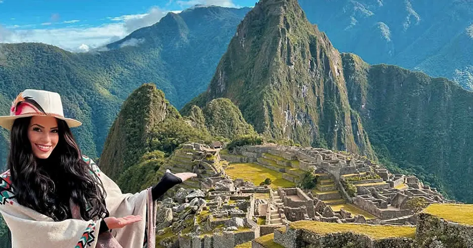 Turista sonriendo mostrando Machu Picchu, con un cielo celeste y rodeada de montañas, en la parte baja se ven los restos arqueológicos de la ciudad inca