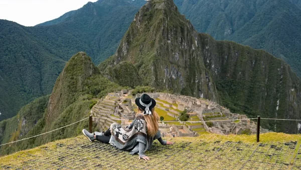 Mujer sentada observando Machu Picchu, en la parte baja los vestigios incas y las grandes montañas alrededor de la ciudadela inca