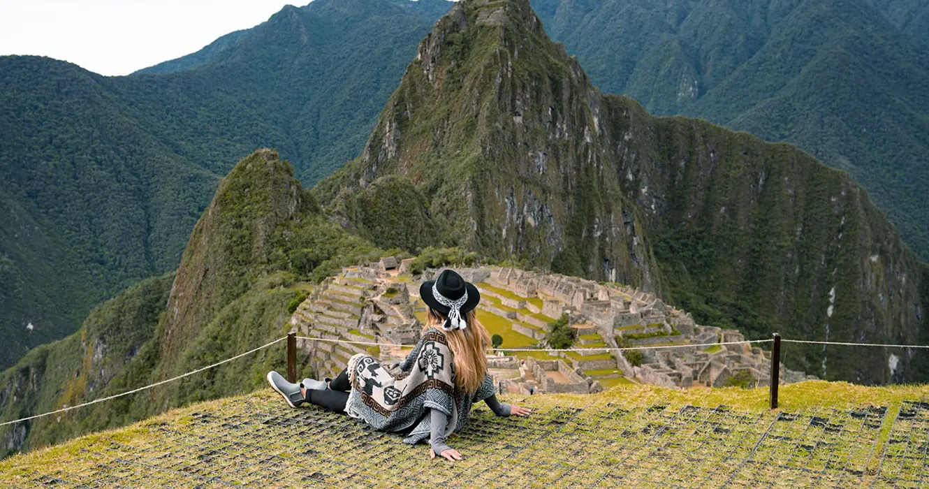 Mujer sentada observando Machu Picchu, en la parte baja los vestigios incas y las grandes montañas alrededor de la ciudadela inca