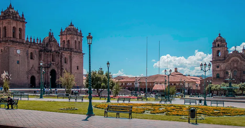Plaza de armas de Cusco con un cielo celeste despejado, bancos en la plaza, se ven las dos iglesias, catedral y compañía de Jesús