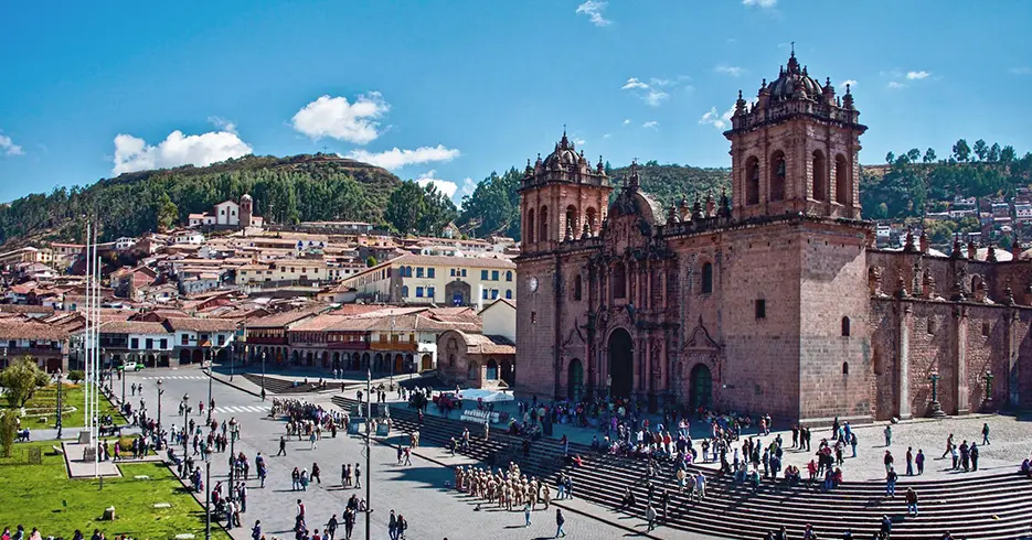 Vista de la catedral de cusco con bastantes personas en la parte baja, en las escaleras y en la calle, árboles verdes al fondo y un cielo celeste
