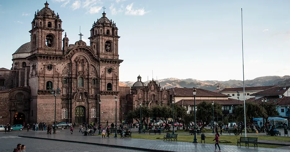 Plaza de armas de cusco con la vista de la iglesia de la compañía de Jesús, varias personas caminando y sentadas en los bancos de la plaza