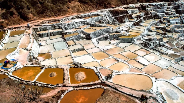Las salineras de maras en cusco, se ven varios pozos de sal, unos con colores blancos, medio beige y marrones, una persona extrayendo la sal de estas piscinas