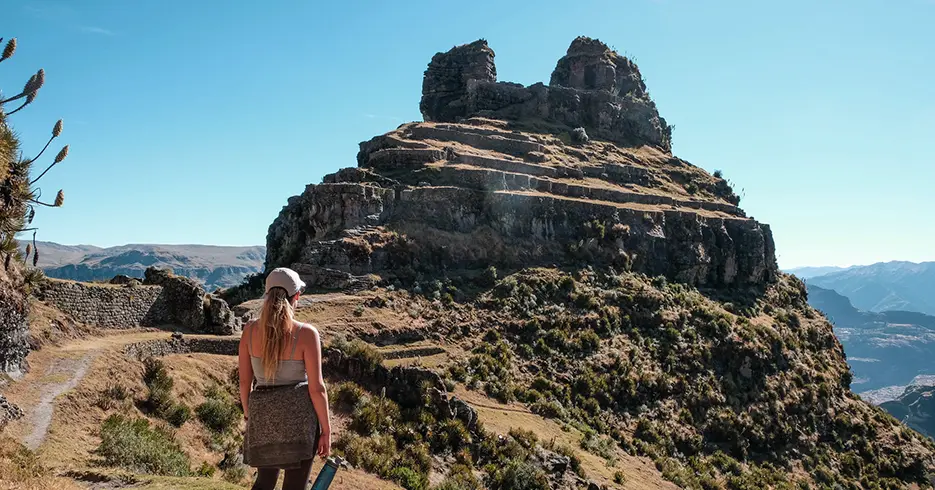 Mujer viendo la fortaleza de waqrapukara en cusco Perú, con un cielo celeste despejado y mucha vegetación alrededor de la fortaleza