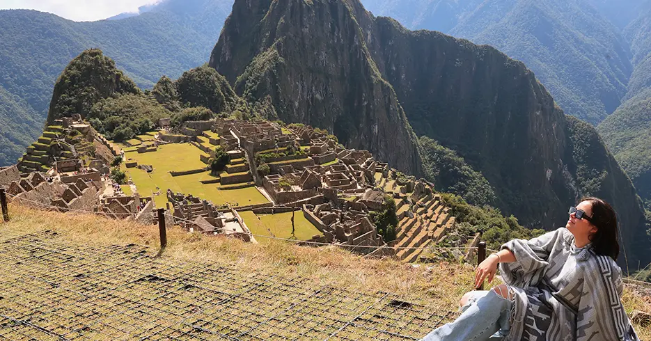 Turista en Machu Picchu sentada, posando para la foto y al fondo en la parte baja se ven los centros arqueológicos con la hermosa vista de las montañas y una vegetación verde