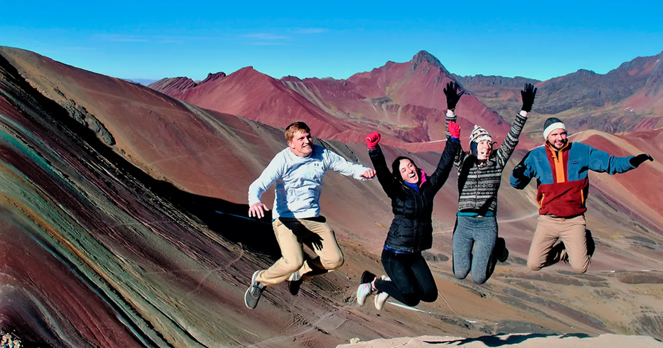 Trekking por la Montaña de los 7 colores: Una Experiencia en familia 10 Turistas saltando en la montaña de los 7 colores, todos felices, al fondo se ven montañas de color verde, rojo y algo de amarillo.